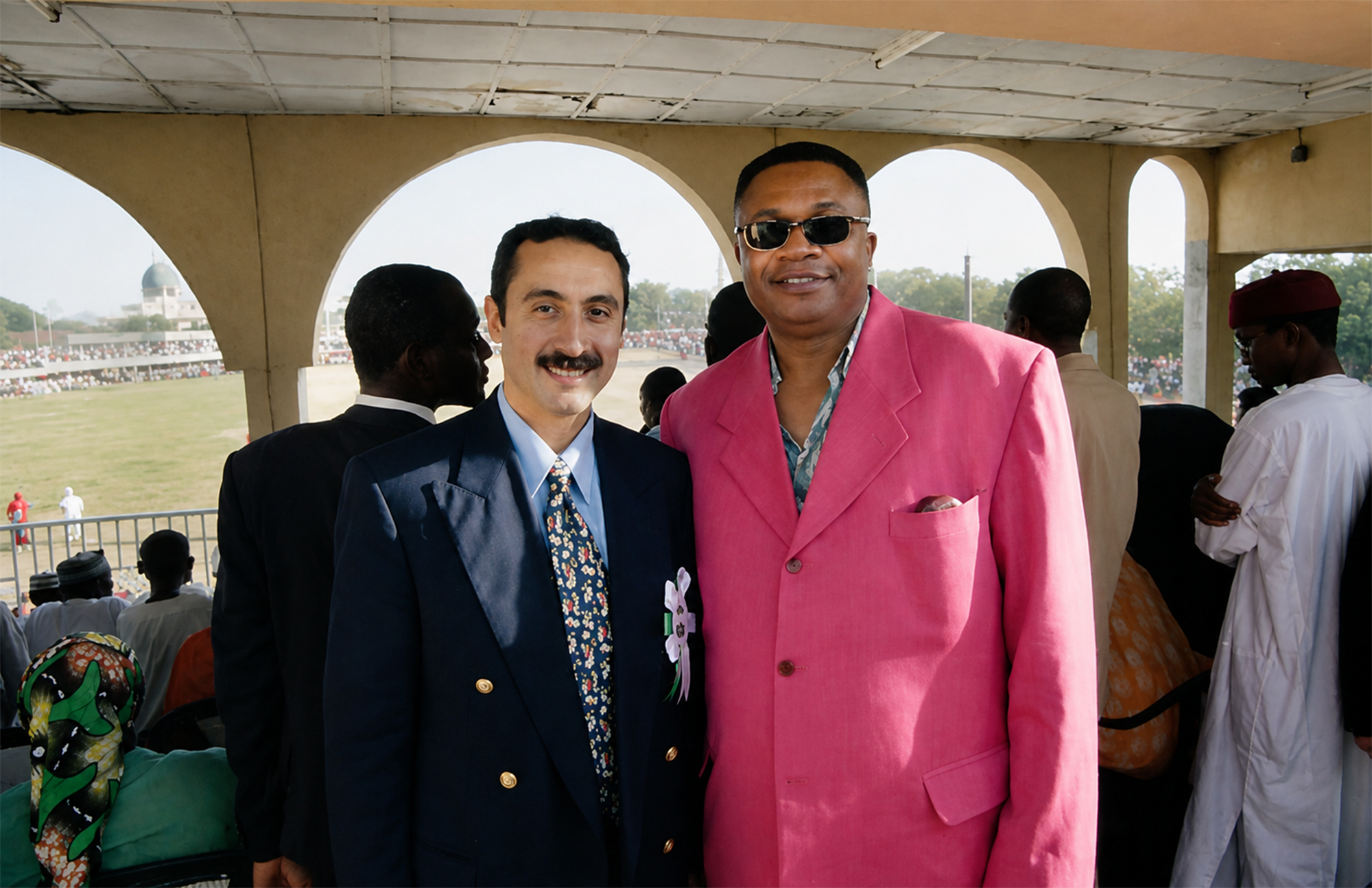 Dos hombres posando bajo una galería con arcos frente a un campo con público durante una ceremonia en Kano durante la Travesía Nigeriana