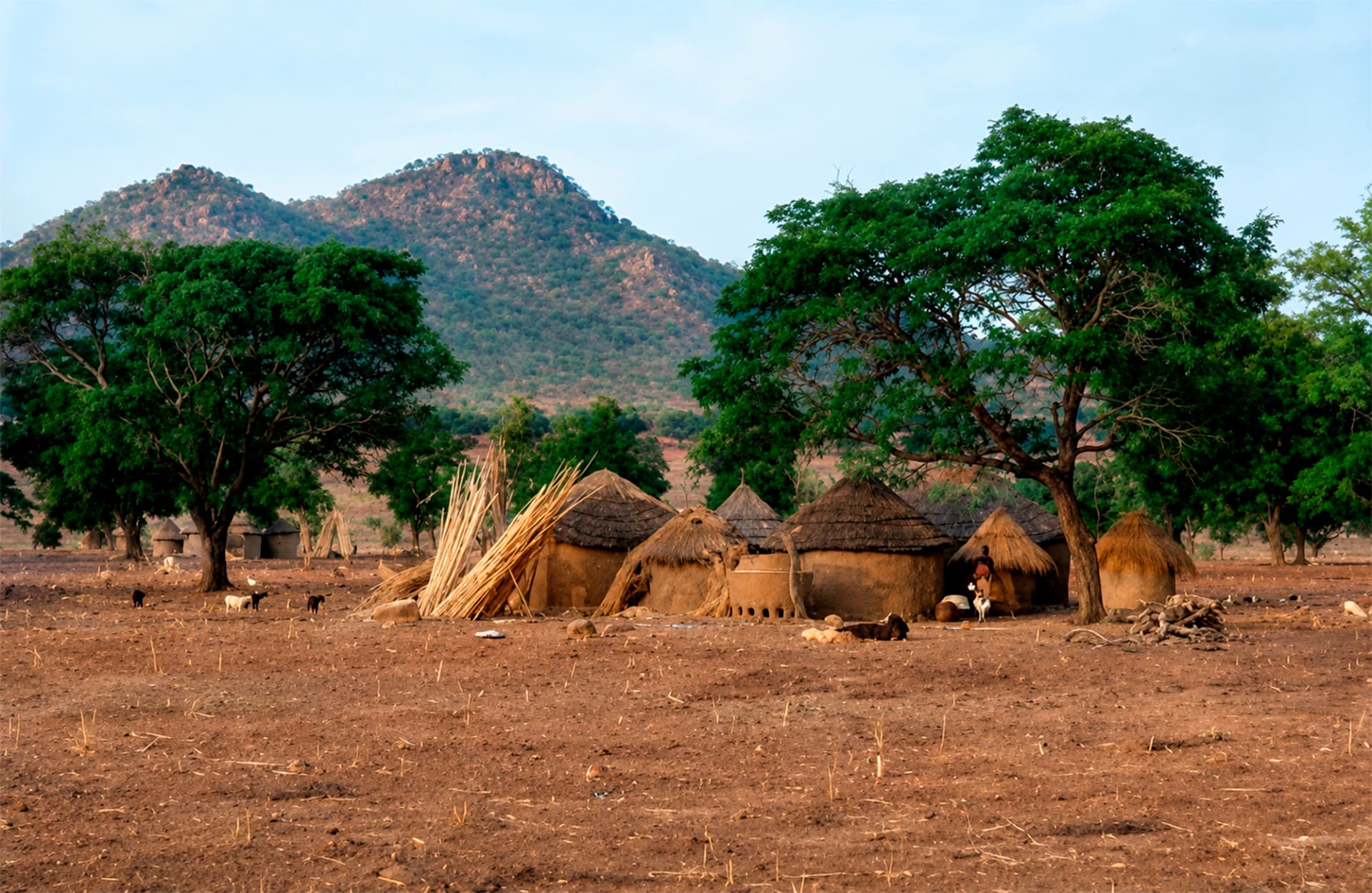 Poblado rural con viviendas circulares de techos de paja, cultivos y montaña rocosa en Jos durante la Travesía Nigeriana