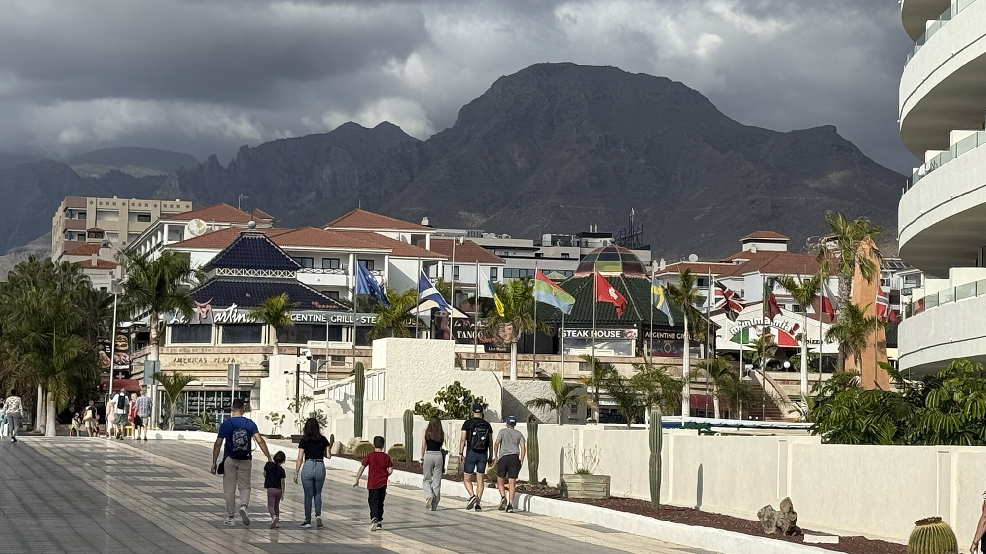 Paseo con palmeras, comercios y banderas ante una montaña escarpada bajo nubes oscuras