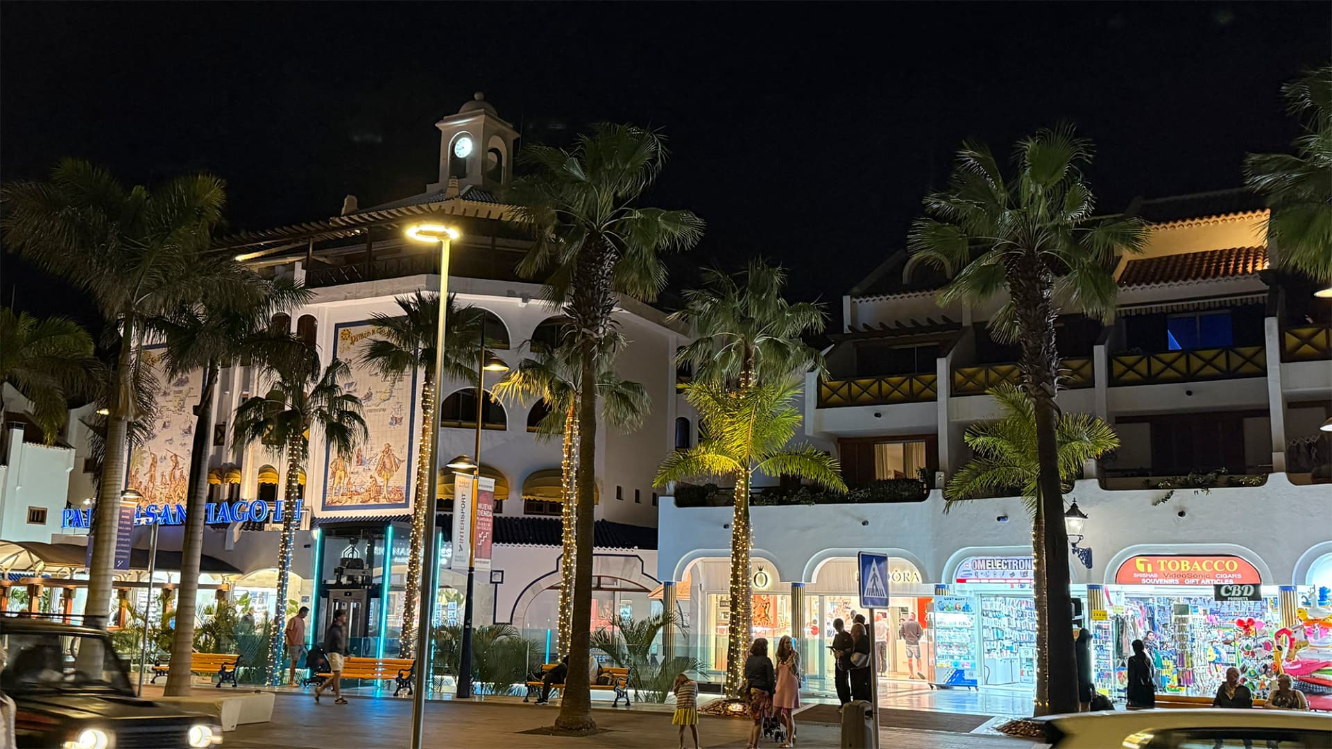 Plaza comercial nocturna con palmeras iluminadas, tiendas y arquitectura blanca