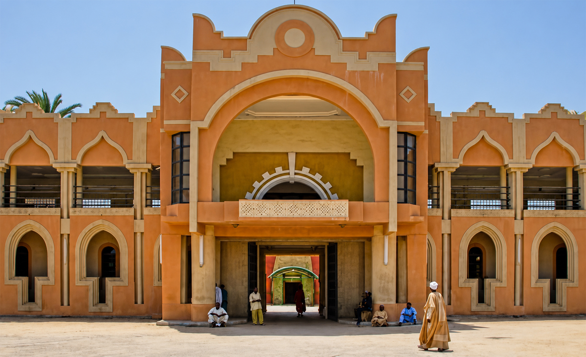 Fachada anaranjada con arcos apuntados y gran portal central, con personas sentadas y un hombre caminando, en Sokoto durante la Travesía Nigeriana